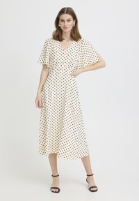 Femme debout portant une robe midi blanche à pois noirs avec des manches volantées, associée à des talons à bride cheville noirs.