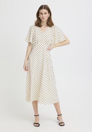 Kvinde står iført en hvid midi-kjole med sorte polkaprikker og flagrende ærmer, kombineret med sorte ankelremssko.
