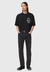 Μαύρο βαμβακερό t-shirt με γραφιστικό σχέδιο στο αριστερό στήθος, συνδυασμένο με σκούρες γκρι τζιν παντελόνι και μαύρες μποτές έως τον αστράγαλο. Μινιμαλιστικό στυλ.