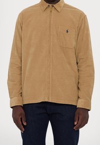 Uomo che indossa una camicia di velluto a coste color cachi con zip e tasca sul petto, e jeans blu scuro, in piedi davanti a uno sfondo neutro.