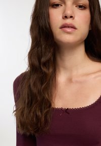 Bordowy top z długim rękawem, z dekoltem w serek, ozdobiony koronkowym wykończeniem i dekoracyjną kokardką na środku. Miękki, gładki materiał.