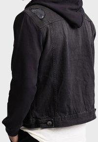 Persoon in een zwarte denim jas met capuchon, zichtbaar is een versleten patch op de schouder en een knoopsluiting aan de zijkant bij de taille.