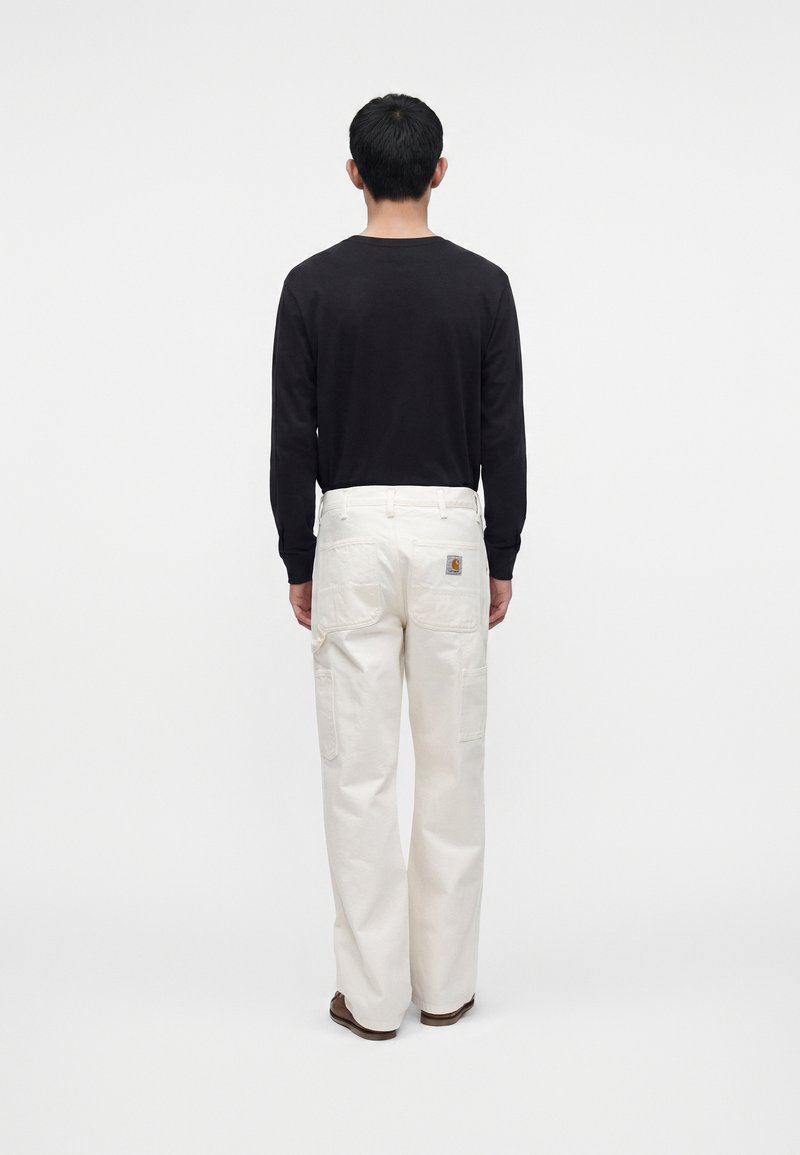 Homme debout, dos tourné, portant une chemise noire à manches longues, un pantalon cargo blanc avec poches arrière et des chaussures marron sur fond blanc uni.