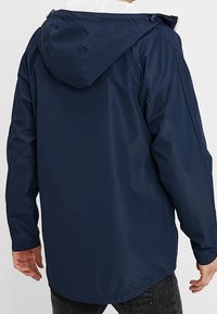 Tummansininen vettä hylkivä takki, jossa on huppu, sileä pinta ja hieman pidempi takaselkä. Ei näkyviä logoja tai kuvioita.