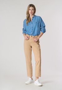Una donna sorridente sta in piedi con sicurezza, indossando una camicia di jeans, pantaloni beige chiari e sneaker bianche, su uno sfondo neutro.