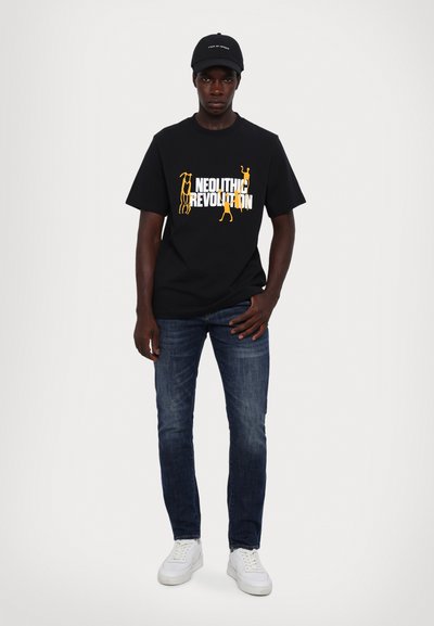 Sort bomulds t-shirt med hvid og orange "Neolitisk Revolution" grafik. Sammen med mørkeblå jeans og hvide sneakers. Sort cap på.