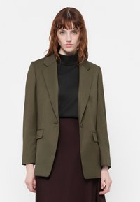 Olivově zelený blazer s přiléhavým střihem, s klopami, jedním knofíkem a dvěma předními kapsami. Nosený přes černý rolák.