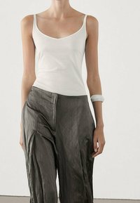 Kobieta ubrana w białą bluzkę bez rękawów, luźne ciemnoszare spodnie z wysokim stanem oraz biały bransolet na lewym nadgarstku.