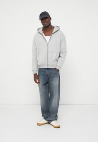 Jauns vīrietis, tērpies pelēkā kapucē, baltā T-kreklā, brīvās zilās džinsās, baltās sporta apavos un melnā beisbola cepurē, stāv pret baltu fona.