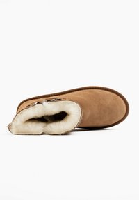 Botín de ante marrón con un suave forro crema, forma redondeada y un acento decorativo de cremallera. Suela plana con un exterior texturizado.