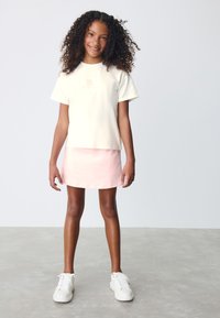 Tricou galben deschis din bumbac cu mâneci scurte și un logo, asortat cu o fustă mini roz, evazată. Purtat cu adidași albi.
