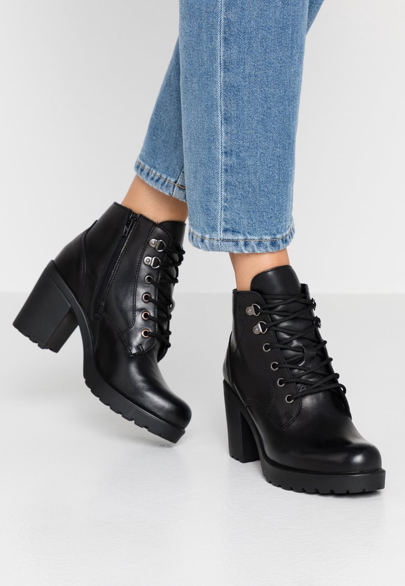 Bottines à talons en cuir noir avec bout arrondi, devant lacé, fermeture éclair et semelle en caoutchouc texturée. Dotées d'accents métalliques œillets.