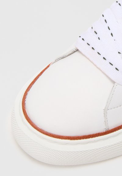 Valge nahast tennis, millel on ümmargune varvas, mille ümber on oranž õmblus ja valged tekstuuriga sneakeri paelad. Minimalistlik disain, lähivõte.