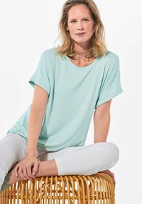 Licht mintgroene t-shirt met korte mouwen, ronde halslijn en subtiel stippenpatroon. Draag het met witte broek, zittend op een geweven stoel.