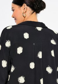 Sort skjorte med store hvide penselstrøg-dots, med krave og rygplissering. Glat tekstur med et løst fit design.
