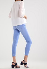 Blusa blanca con mangas transparentes, jeans skinny azules y sandalias de tacón cuadrado negras. Enfoque en líneas limpias y diseño contemporáneo.