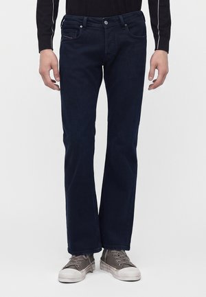 Person iført mørkeblå jeans med lige ben og grå afslappede sneakers, der står mod en ensfarvet baggrund.