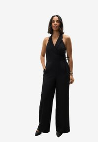 Jumpsuit noir à col halter avec un design croisé, une silhouette à jambes larges, des poches latérales et un détail à nouer à la taille. Tissu lisse et léger.