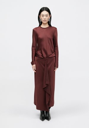Duga burgundska haljina s dugim rukavima i uvijenim dizajnom u struku, uparena s crnim čizmama na petu. Glatka tekstura, minimalistički stil.