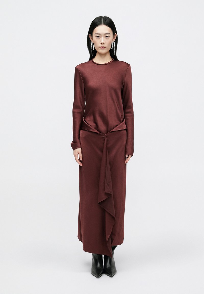 Vestido longo burgundy com mangas longas e um design torcido na cintura, combinado com botas pretas de salto. Textura suave, estilo minimalista.