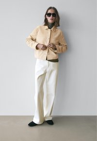 Beige ruskindsjakke med krave, frontknapper og sidelommer, kombineret med off-white vidde bukser og sorte slip-on sko.