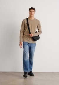 Beige lang mouw shirt, ontspannen pasvorm blauwe spijkerbroek en zwarte sneakers. Zwarte crossbody tas. Neutrale achtergrond, onderwerp aan het lopen.