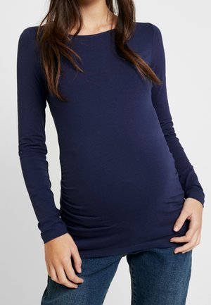 Femme portant une chemise bleu marine à manches longues et un jean bleu, tenant son ventre de femme enceinte avec les deux mains.