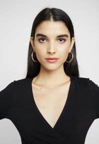 Top preto envolvente com decote em V profundo, mangas curtas e textura suave. O modelo usa grandes brincos de argola dourados. Fundo neutro.