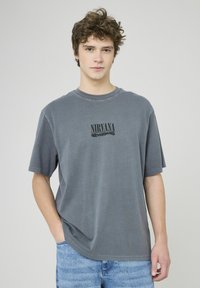 T-shirt gris surdimensionné en coton à manches courtes. Présente un logo noir "NIRVANA" imprimé au centre. Ajustement décontracté, texture lisse.