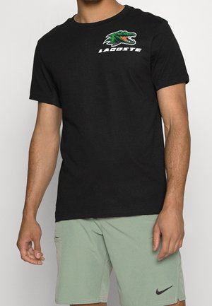 Man draagt een zwart Lacoste T-shirt met groen krokodillenlogo en lichtgroene Nike shorts tegen een effen achtergrond.