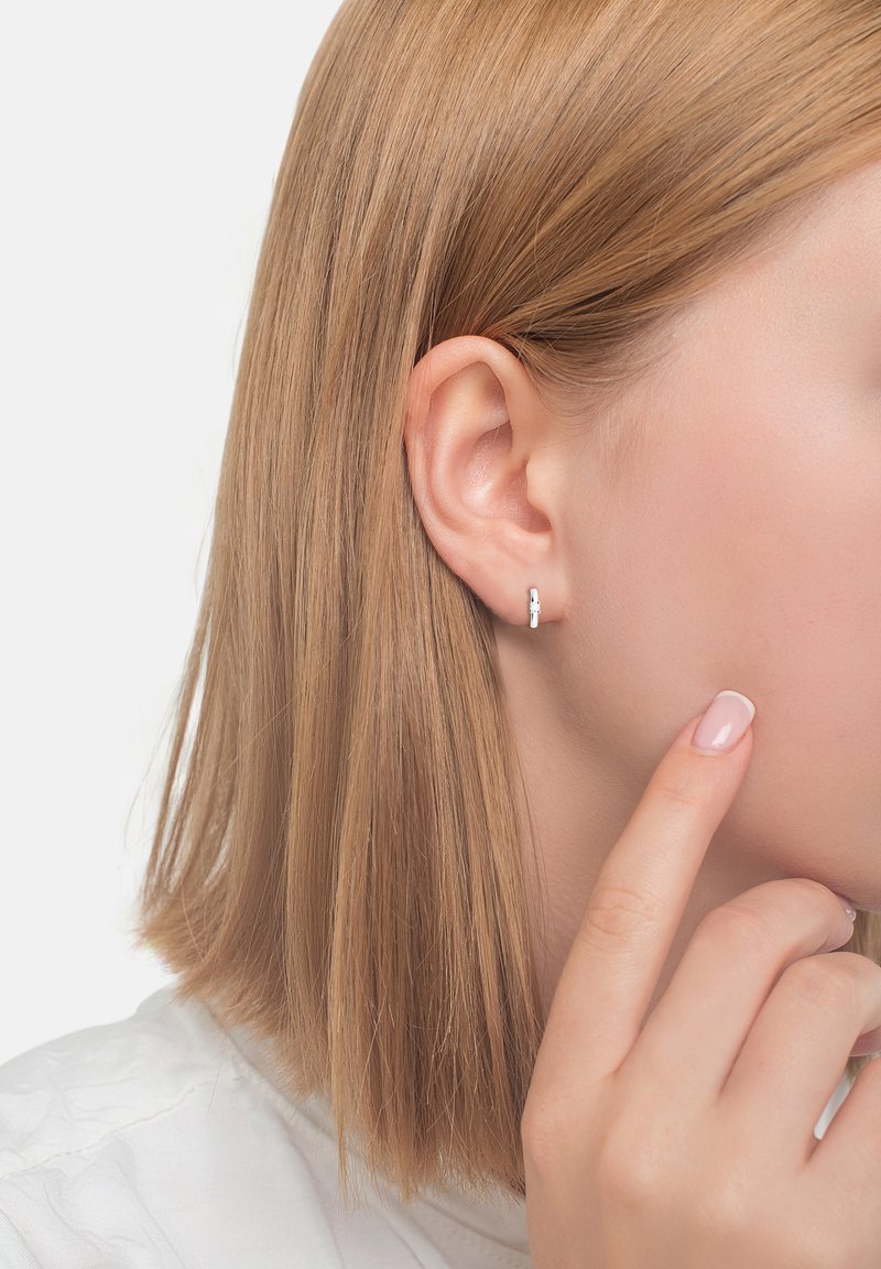 Silberne Creolen, die am Ohrläppchen getragen werden, mit einer glatten Oberfläche und einem schlichten Design. Das Haar ist glatt und hellblond, vor einem neutralen Hintergrund.