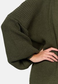 Olivgrön stickad cardigan med ett texturerat mönster, översized ärmar och ribbade muddar. Tyget verkar mjukt och tjockt.