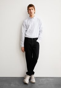 Chemise à rayures bleu clair avec du blanc, associée à un jean noir et des baskets beiges. Pose debout sur un fond uni.