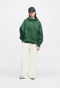 Person som har på seg en grønn Nike-hettegenser, hvite Nike joggebukser, svart Nike caps og hvite joggesko, står mot en ensfarget hvit bakgrunn.