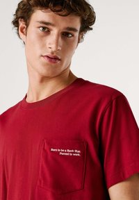 Rood katoenen T-shirt met een ronde hals en korte mouwen. Heeft een borstzakje met witte tekst: "Born to be a Rock Star. Forced to work."