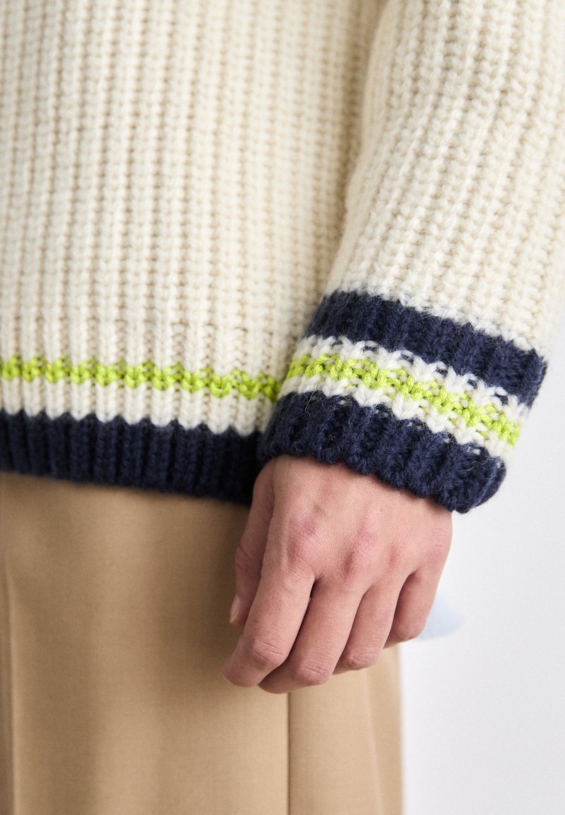 Pull en maille chunky crème, avec des rayures bleu marine et vert lime au niveau des poignets. Tissu texturé avec des détails en côtes.