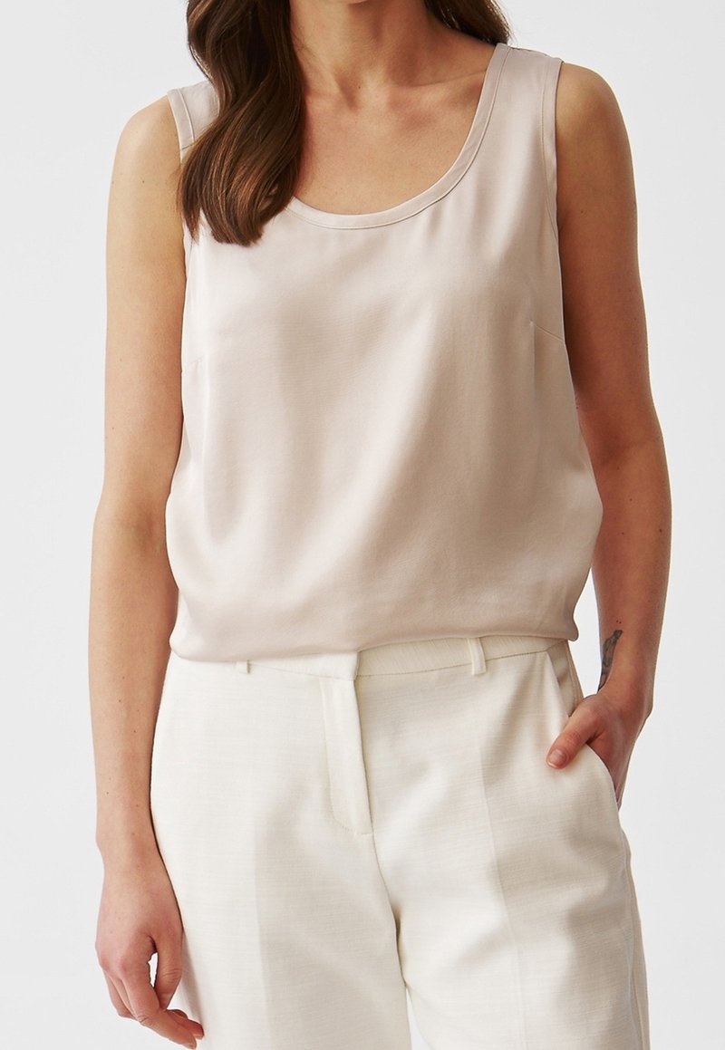 Femme portant un haut beige clair sans manches rentré dans un pantalon crème taille haute, avec une main dans la poche et de longs cheveux bruns sur l'épaule.