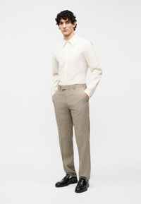 Άνδρας με σγουρά μαλλιά που φοράει εκρού πουκάμισο με κουμπιά, μπεζ παντελόνι και μαύρα παπούτσια, στέκεται με τα χέρια στις τσέπες.