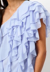 Lichtblauwe top met gelaagde ruffles op één schouder, gemaakt van een zachte, lichte stof. Gladde textuur met een vloeiende uitstraling.