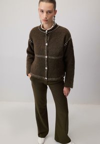 Femeie în picioare cu mâinile în buzunare, purtând o cardigană călduroasă maro cu nasturi, pantaloni verzi măslinii, încălțăminte albă și cercei argintii atârnători.