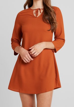 Femme portant une robe à manches longues couleur rouille avec un col en trou de serrure noué par un nœud, debout devant un fond uni.