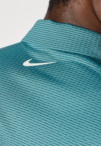 Τυρκουάζ πόλο μπλουζάκι με υφή, με λευκό λογότυπο Nike στον γιακά. Το ύφασμα φαίνεται ελαφρύ και αναπνεύσιμο.