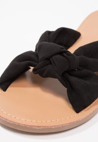 Sandale plate avec semelle couleur tan et larges brides en tissu noir nouées en un grand nœud sur la zone des orteils.