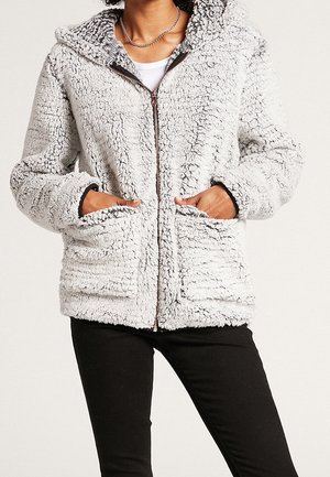 Osoba ubrana w jasnoszarą, miękką kurtkę na zamek z przednimi kieszeniami i kapturem, w połączeniu z czarnymi spodniami i białą koszulką pod spodem.