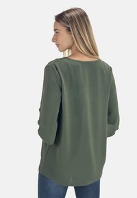 Olijfgroene blouse met driekwart mouwen, relaxed fit, textuurstof en een ronde halslijn, gecombineerd met denim broek.