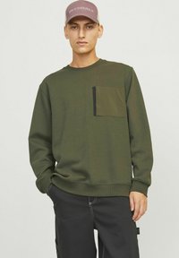 Olivenfarbener Pullover mit einer Fronttasche in einem kontrastierenden Dunkelgrün, runder Ausschnitt und gerippten Bündchen, kombiniert mit schwarzen Hosen.