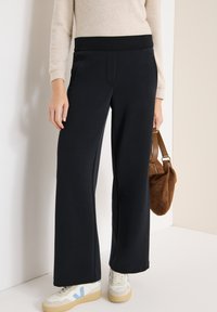 Pantalon de survêtement noir à jambes larges avec une taille élastique, des poches latérales et une texture lisse, associé à des baskets blanches et un sac marron.