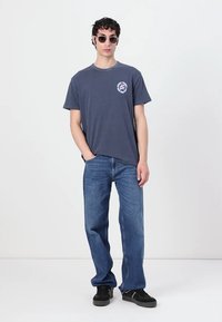 Mørk blå bomull T-skjorte med et sirkulært grafisk motiv på venstre bryst, kombinert med lyseblå denimjeans og svarte joggesko.