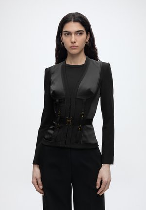 Woman with long dark hair wears black fitted blazer with structured leather corset and black pants, standing against a plain white background.