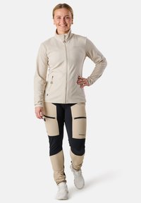 Veste en polaire beige avec fermeture éclair devant et poches latérales, associée à un pantalon beige et noir bicolore doté de poches zippées sur les cuisses.